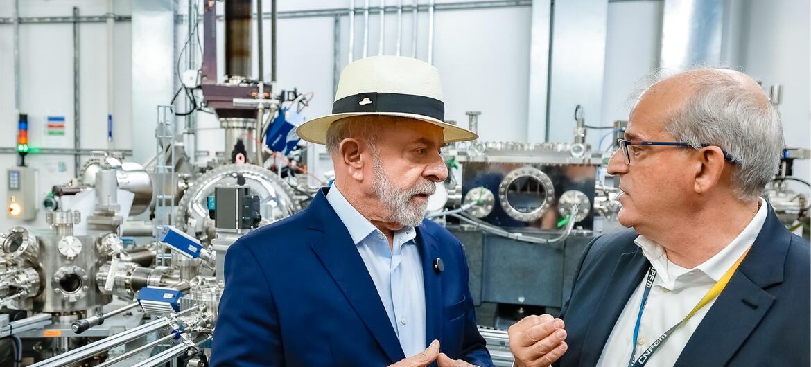 Lula da Silva, usando um chapéu e um terno azul, está conversando com outro homem de terno escuro dentro de uma instalação industrial de alta tecnologia.