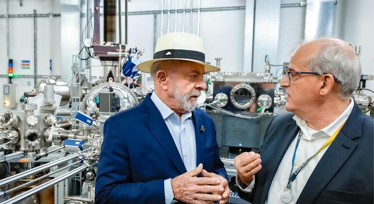 Lula da Silva, usando um chapéu e um terno azul, está conversando com outro homem de terno escuro dentro de uma instalação industrial de alta tecnologia.