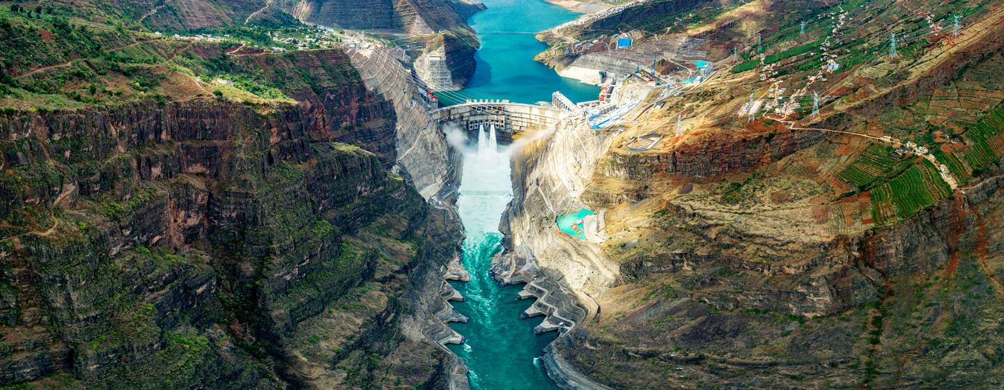 Vista aérea de una enorme presa hidroeléctrica incrustada en un profundo cañón de montaña con agua turquesa que fluye a través, mostrando la infraestructura de energía limpia.