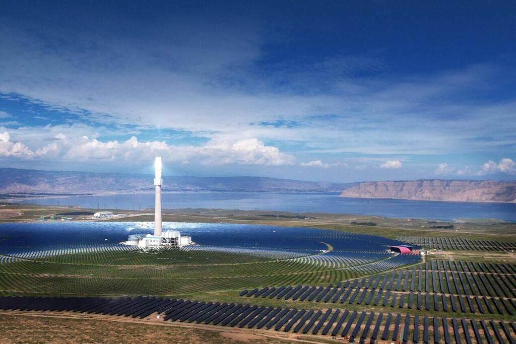 Une centrale solaire étendue avec des rangées de panneaux solaires et une tour solaire centrale émettant de la lumière, sur fond d'un grand lac et de montagnes éloignées sous un ciel partiellement nuageux.