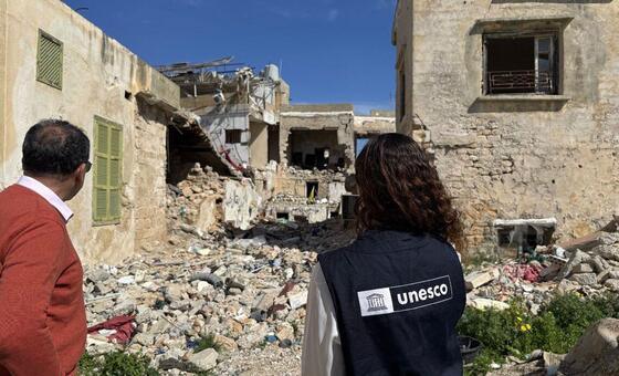Two people, a man in a red sweater and a woman wearing a UNESCO vest, stand looking at destroyed buildings and rubble in the Middle East. The scene depicts the aftermath of violence, with UNESCO providing emergency support to protect education, culture, and journalists.