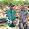 Sehemu ya mahojiano ya nje huko Mwanza, Tanzania, akishirikiana na mwandishi akishika kipaza sauti na kuzungumza na mtu wa eneo hilo. Wanaketi kwenye pwani wakiwa wamezungukwa na mashua nyingi za uvuvi za mbao zenye rangi na nyavu za uvuvi, na mwili mkubwa wa maji unaonekana nyuma.