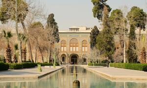 A wide shot of the Golestan Palace in Iran, featuring its ornate tiled facade reflected in a long rectangular pool of water, surrounded by manicured gardens and trees.