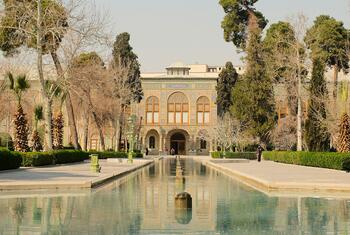 A wide shot of the Golestan Palace in Iran, featuring its ornate tiled facade reflected in a long rectangular pool of water, surrounded by manicured gardens and trees.