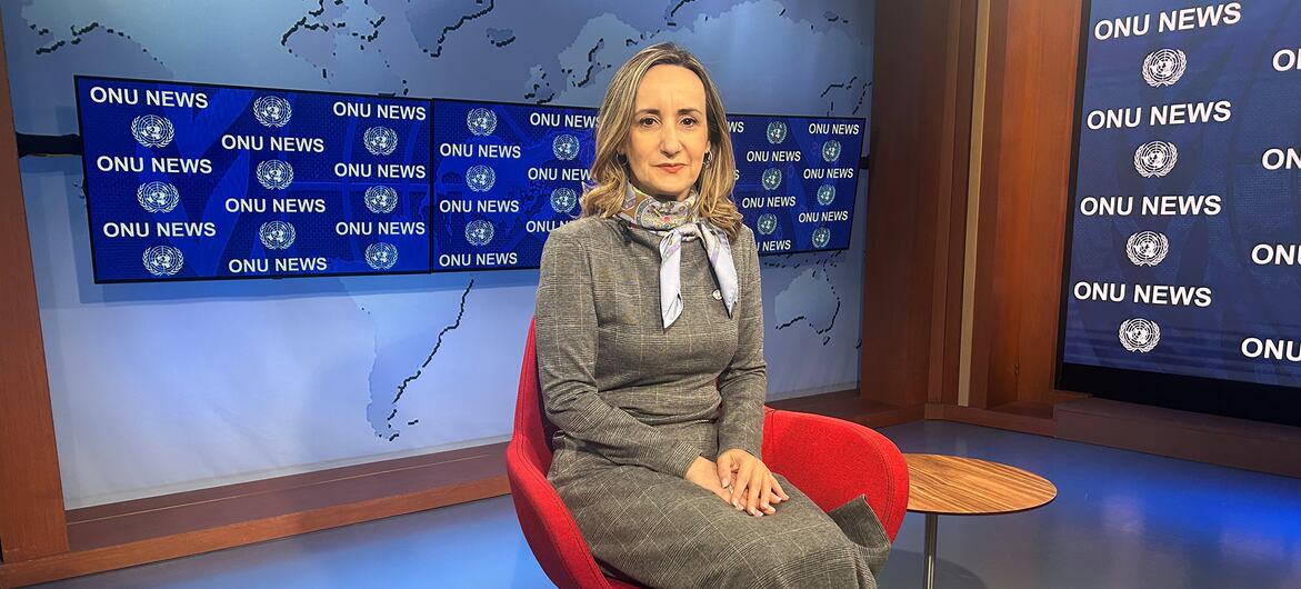 Uma apresentadora de notícias vestindo um terno cinza está sentada em uma cadeira vermelha em um estúdio de televisão. O fundo apresenta um mapa-múndi e telas exibindo o logotipo "ONU NEWS".