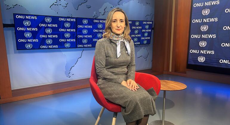 Uma apresentadora de notícias vestindo um terno cinza está sentada em uma cadeira vermelha em um estúdio de televisão. O fundo apresenta um mapa-múndi e telas exibindo o logotipo "ONU NEWS".