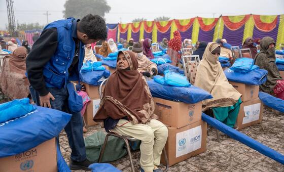 Os sobreviventes das inundações no Paquistão recebem ajuda em um acampamento de socorro, com famílias deslocadas sentadas em caixas de papelão ao lado de seus pacotes de ajuda azuis, enquanto um jovem vestindo um colete azul da OIM fala com uma mulher usando um xale marrom. A cena destaca as consequências das inundações do rio Chenab em Punjab.