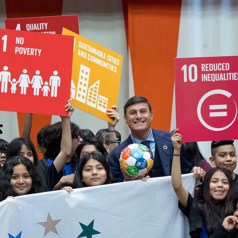 Javier Zanetti holds a soccer ball while surrounded by children holding signs for the United Nations Sustainable Development Goals at an event.
