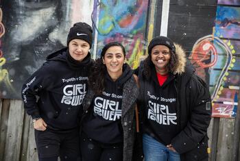 Three women wearing 'Girl Power' hoodies stand together in front of a graffiti-covered wall.