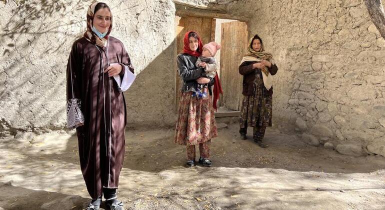Три афганских женщины, в том числе матери, содержащие младенцев, стоят за традиционным кирпичным домом в сельской местности, подчеркивая усилия Всемирной продовольственной программы по оказанию гуманитарной помощи.