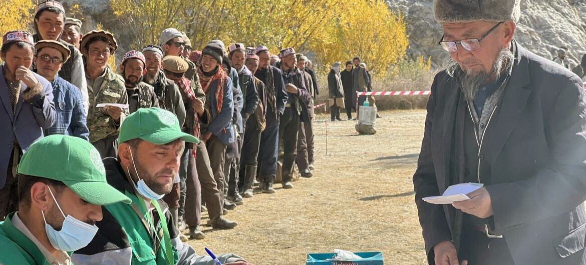 Um trabalhador humanitário do Programa Mundial de Alimentos, vestido de colete verde e chapéu, assina documentos numa mesa, enquanto um afegão local com um fato escuro e óculos recebe papéis.