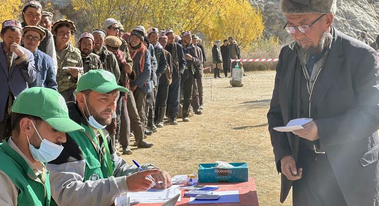 ग्रीन जैकेट और टोपी पहने विश्व खाद्य कार्यक्रम के एक सहायता कार्यकर्ता ने एक मेज पर दस्तावेजों पर हस्ताक्षर किए, जबकि अंधेरे सूट और चश्मे में एक स्थानीय अफगान व्यक्ति को कागजात प्राप्त हुए। ग्रामीण बाहरी वातावरण में लोगों की लंबी कतार उनके पीछे इंतजार कर रही है।