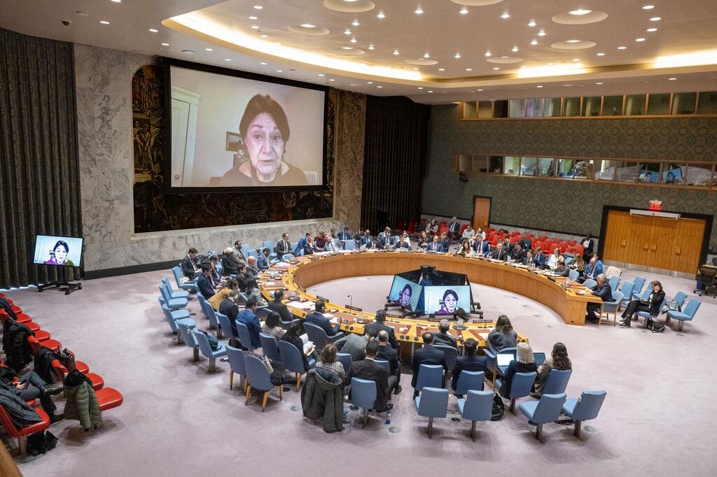 A wide shot of the United Nations Security Council chamber during a non-proliferation meeting, with delegates seated around a large circular table and a large screen displaying a speaker participating via video conference.