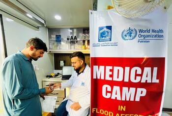 पंजाब, पाकिस्तान में बाढ़ प्रभावित समुदायों को स्वास्थ्य सेवाएं प्रदान करने वाला डब्ल्यूएचओ का एक मोबाइल चिकित्सा शिविर, जिसमें एक स्वास्थ्य कार्यकर्ता एक रोगी की सहायता कर रहा है।
