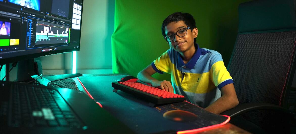 Raul John Aju, un jeune de 16 ans portant des lunettes et un polo à rayures jaune et bleu, sourit à la caméra alors qu'il est assis à un bureau d'ordinateur avec un grand moniteur affichant un logiciel de montage vidéo et un fond d'écran vert.