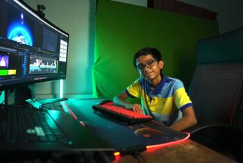 Raul John Aju, a 16-year-old wearing glasses and a yellow and blue striped polo shirt smiles at the camera while sitting at a computer desk with a large monitor displaying video editing software and a green screen backdrop.