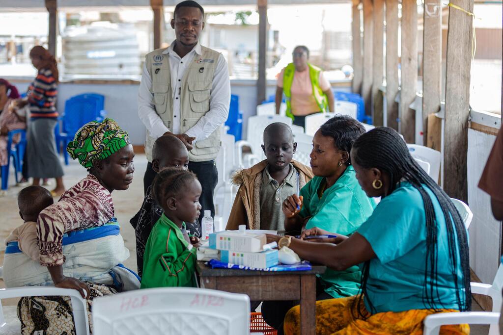 Une famille avec des enfants reçoit des soins médicaux dans un établissement du HCR à Uvira, en République démocratique du Congo. Un membre du personnel du HCR vêtu d'un gilet beige se tient derrière le groupe, tandis que deux travailleuses de la santé en robes vertes s'occupent des patients à une table.