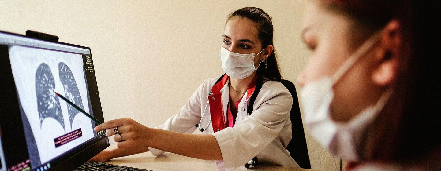 Une femme médecin portant un masque montre une radiographie thoracique des poumons sur un moniteur d'ordinateur alors qu'elle parle à un patient aussi masqué.