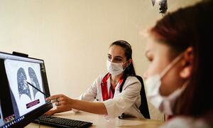 A female doctor wearing a mask points to a chest X-ray of lungs on a computer monitor while speaking to a patient also wearing a mask.