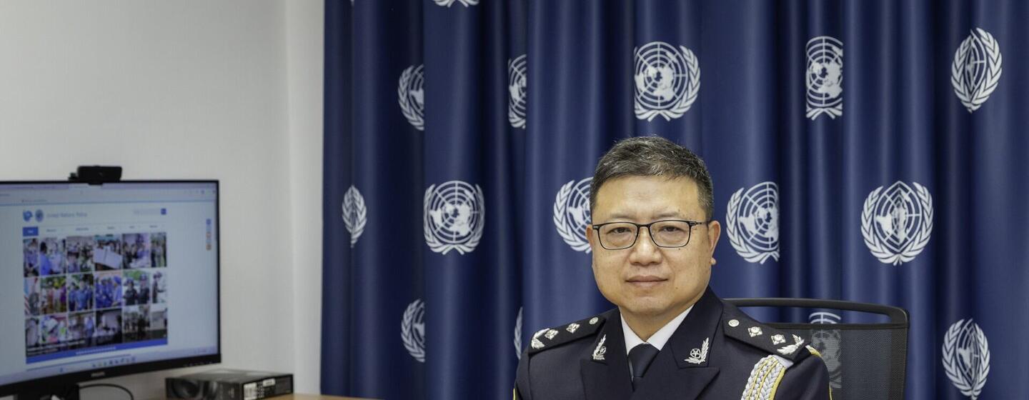 一个穿着中国军装的男子坐在办公室的办公桌上,背景是联合国旗.