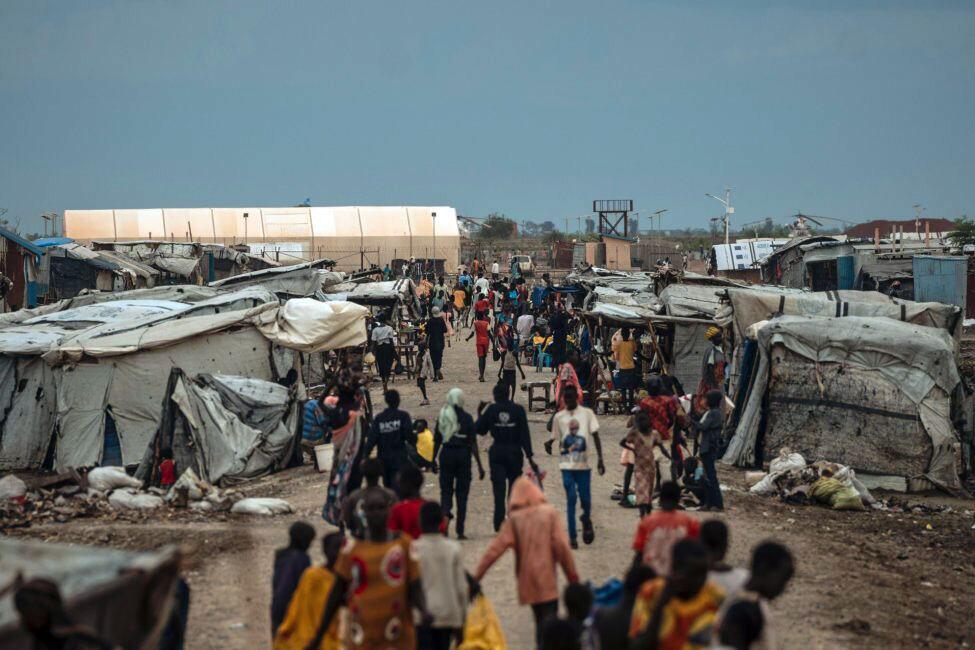 Njia iliyojaa watu kupitia kambi ya muda huko Sudan Kusini, na watu waliohamishwa wakitembea kati ya makazi ya vitambaa chini ya anga safi.