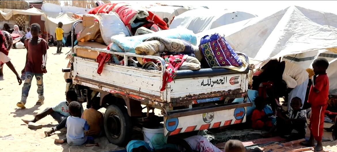 تتحرك شاحنة محملة بأغراض شخصية عبر مخيم للنازحين في غوز السلام، السودان، محاطة بالأطفال والنساء. يعكس هذا المشهد الأزمة الإنسانية والنزوح الناجم عن النزاع في منطقة النيل الأبيض.