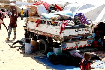 تتحرك شاحنة محملة بأغراض شخصية عبر مخيم للنازحين في غوز السلام، السودان، محاطة بالأطفال والنساء. يعكس هذا المشهد الأزمة الإنسانية والنزوح الناجم عن النزاع في منطقة النيل الأبيض.