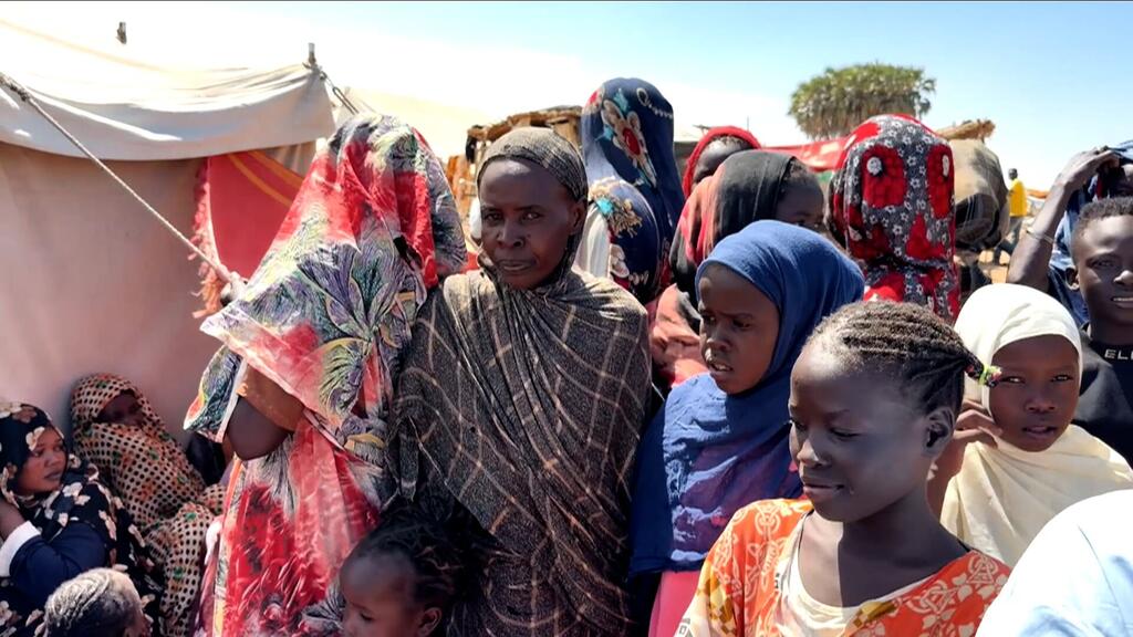 مجموعة من النساء والأطفال السودانيات، كثير منهم يرتدون حجابات ملونة، يقفون في مخيم للنازحين تحت سماء صافية، مع خيام مرئية في الخلفية.