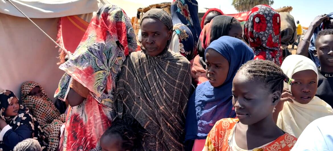 A group of Sudanese women and children, many wearing colorful headscarves, stand in a displacement camp under a clear sky, with tents visible in the background.