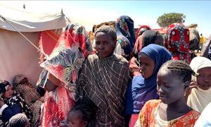 A group of Sudanese women and children, many wearing colorful headscarves, stand in a displacement camp under a clear sky, with tents visible in the background.