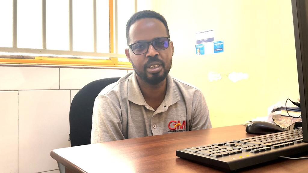 Picha ya mtu mweusi mwenye miwani na ndevu, amevaa shati ya rangi ya kijivu yenye nembo ya 'GM', ameketi kwenye dawati la mbao katika ofisi.