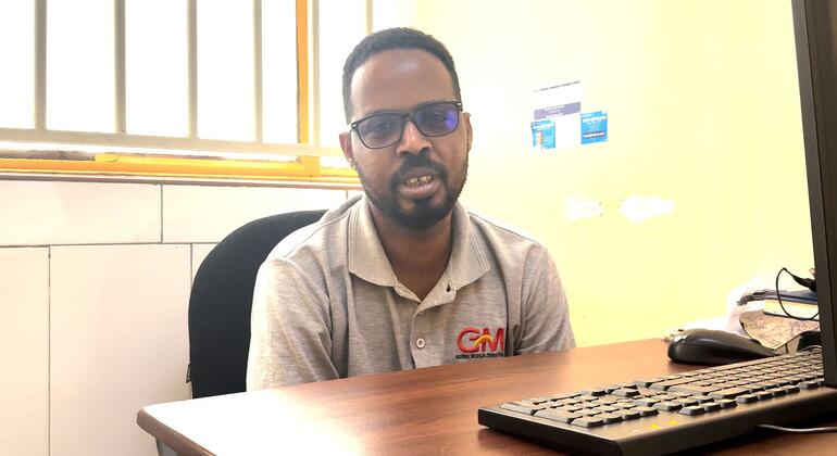 صورة للرجل الأسود ذو النظارات واللحية، يرتدي قميص البولو الرمادي مع شعار "GM"، يجلس على مكتب خشبي في مكتب.