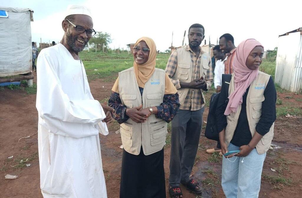 Mwanamume Mwafrika mwenye kanzu nyeupe na kofia anashikana mikono na wafanyakazi wawili wa kike wa misaada wakiwa wamevaa mavazi ya MDPD na hijab katika mazingira ya vijijini.