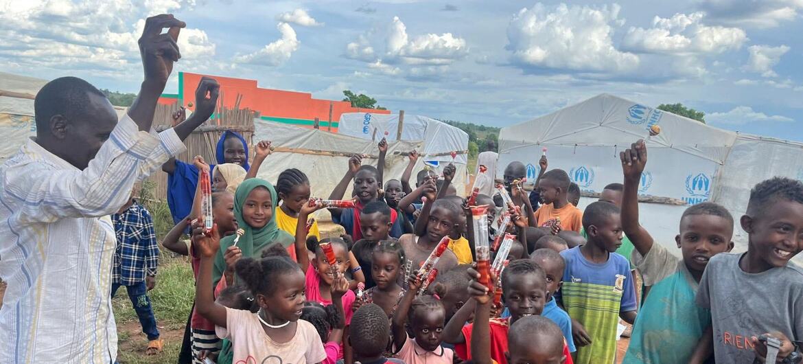 A group of smiling African children in a refugee camp in Uganda, holding up bars of soap, with a man distributing aid in the background.