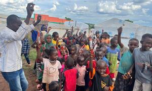 A group of smiling African children in a refugee camp in Uganda, holding up bars of soap, with a man distributing aid in the background.