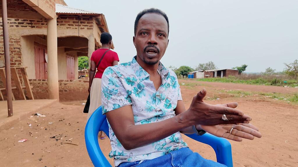 Mwanamume mwenye ndevu na nywele fupi, amevaa shati la maua, ameketi kwenye kiti cha bluu na kufanya ishara wakati akizungumza. Nyuma yake, mwanamke mwenye shati nyekundu anatembea karibu na jengo la matofali katika mazingira ya vijijini.