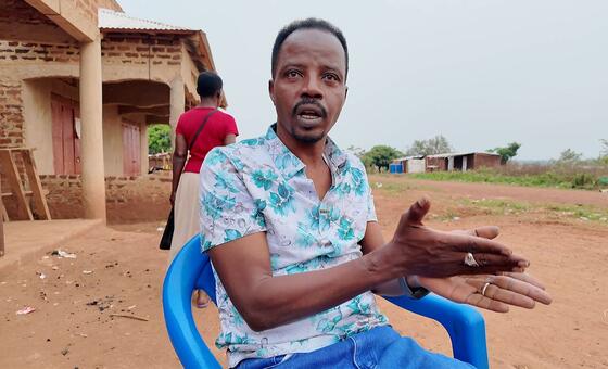 رجل ذو شارب وشعر قصير، يرتدي قميصًا زهريًا، يجلس على كرسي أزرق ويتحرك أثناء الكلام. وراءه، تمشي امرأة في قميص أحمر أمام مبنى من الطوب في بيئة ريفية.