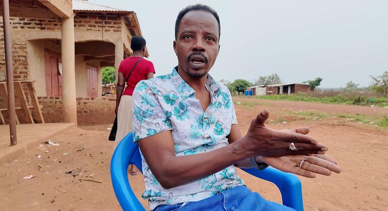 За ним женщина в красной рубашке проходит мимо кирпичного здания в сельской местности.