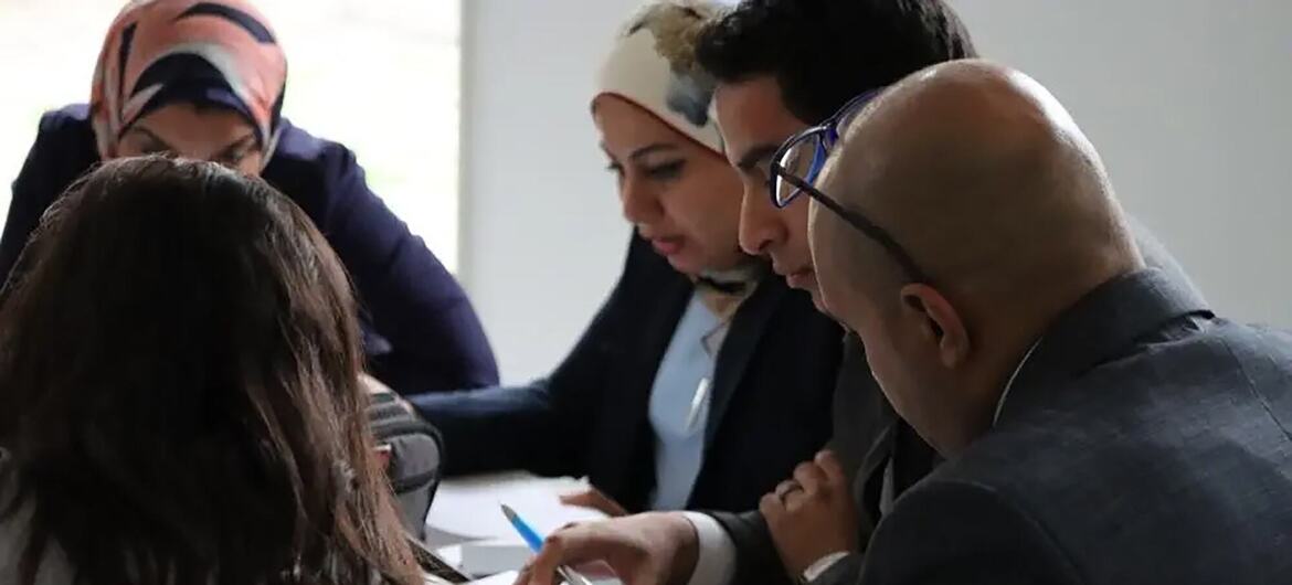 Um grupo diversificado de profissionais vestidos de negócios reúne-se em torno de uma mesa numa sala de reuniões, revisando documentos e discutindo estratégias.