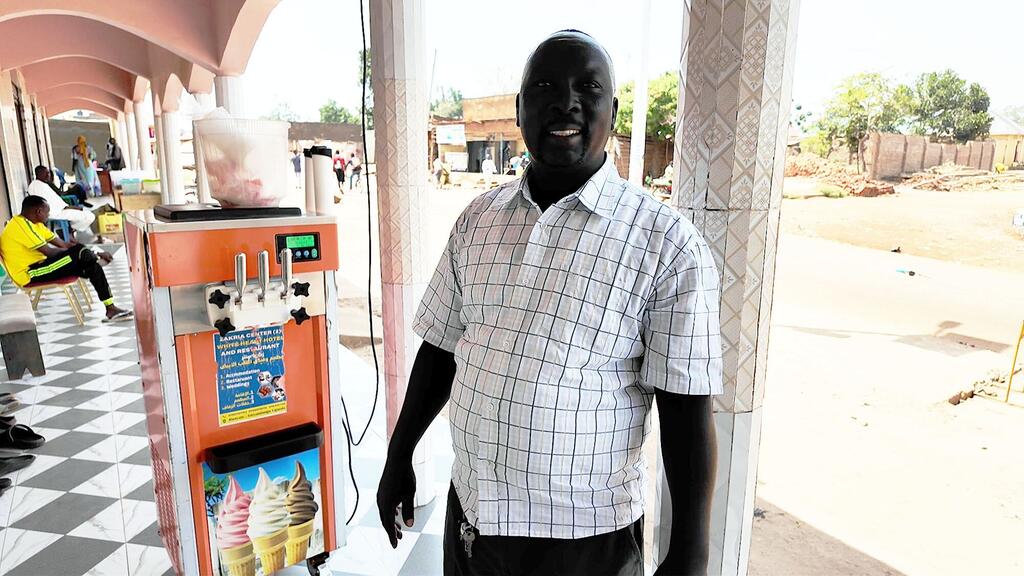 Mwanamume mwenye tabasamu, Ibrahim Zakaria Yahya, amesimama kando ya mashine ya saizi ya barafu katika duka nchini Uganda.