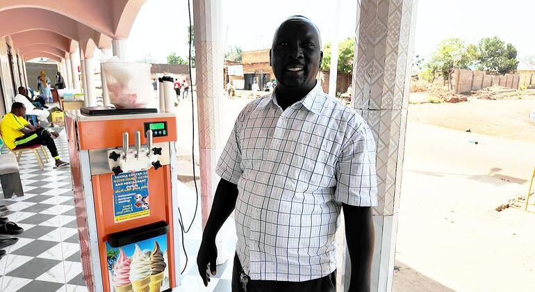 رجل مبتسم، إبراهيم زكريا يحيى، يقف بجانب آلة الآيس كريم في متجر في أوغندا.
