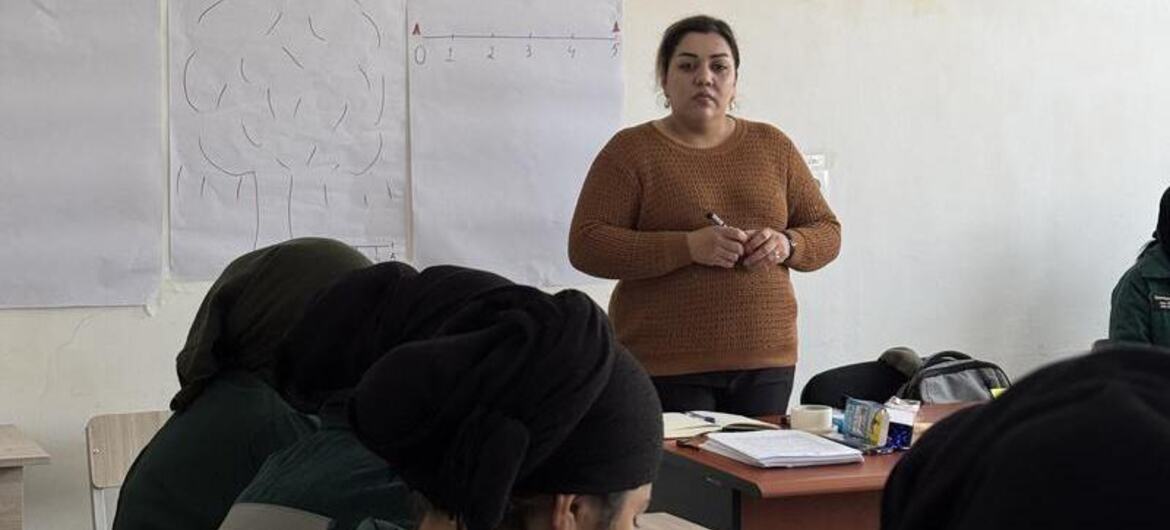 Uma instrutora está na frente de uma sala de aula no Tajiquistão, ensinando a um grupo de alunos que estão sentados em mesas. Um quadro com diagramas é visível atrás dela.