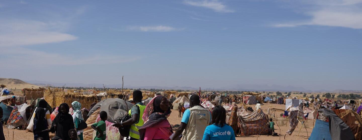 Un travailleur de l'UNICEF en chemise bleue traverse un camp de réfugiés dans le désert du Soudan, entouré de familles déplacées et de refuges provisoires.