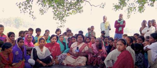 Un grupo de mujeres reporteras y miembros de la comunidad se reunieron al aire libre bajo árboles, participando en discusiones o informes, representando a los medios de comunicación de base en la India.