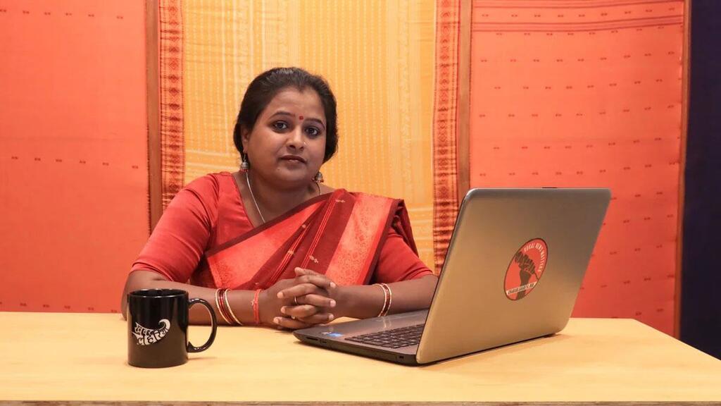 Una mujer reportera vestida de sari rojo se sienta en un escritorio con una computadora portátil y una taza, simbolizando los medios de comunicación de base en la India cambiando las narrativas sobre temas de género.