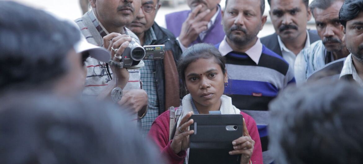 एक युवा भारतीय महिला रिपोर्टर एक टैबलेट रखती है, जो पुरुषों के एक समूह के आसपास है, जो भारत में मीडिया की कथाओं को बदलने में महिलाओं की भूमिका पर प्रकाश डालती है।