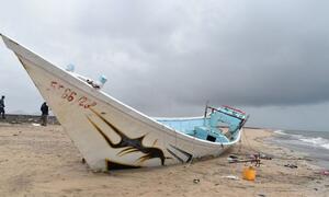 A damaged white and blue boat washed ashore on a beach in Djibouti, with two people standing in the background under a cloudy sky.