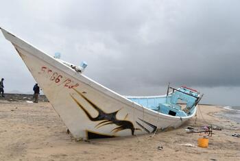 Un bateau blanc et bleu endommagé a débarqué sur une plage de Djibouti, avec deux personnes debout en arrière-plan sous un ciel nuageux.