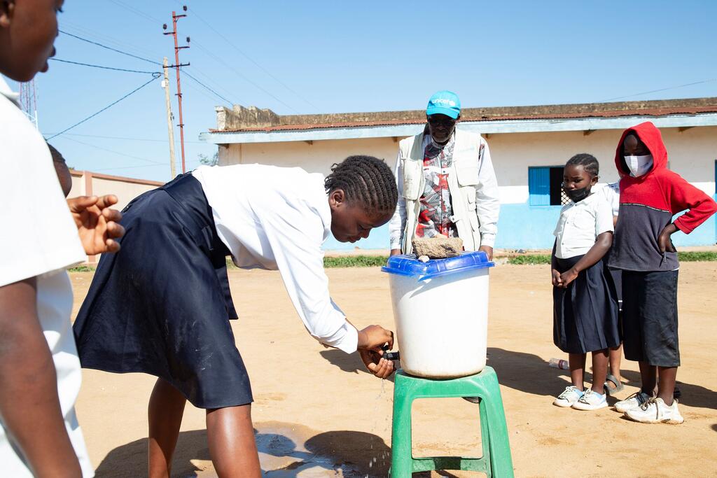 Msichana mchanga aliyevaa sare ya shule anaosha mikono yake kwenye chombo cha maji nje ya shule moja nchini DR Congo, huku mwanamume aliyevaa koti la UNICEF na wanafunzi wengine wakitazama.