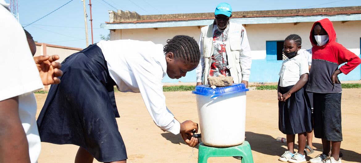 Une jeune fille en uniforme scolaire se lave les mains à un récipient d'eau à l'extérieur d'une école en RDC, tandis qu'un homme portant un gilet de l'UNICEF et d'autres élèves regardent.